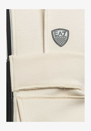 Tessuto sportivo beige con finitura strutturata, caratterizzato da una zip nera e un logo a scudo. Il design include un dettaglio di tasca orizzontale.