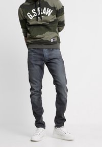 Mikina s kamuflážovým vzorem v tmavě zelené barvě s bílým logem; slim-fit šedé džíny; bílé tenisky. Neformální městské oblečení s kontrastními barvami.