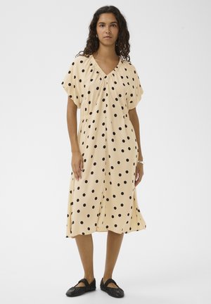 Femme aux cheveux bouclés portant une robe midi beige à pois noirs et des chaussures plates noires, debout devant un fond uni.