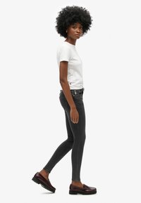 T-shirt blanc ajusté, jeans skinny foncés et mocassins bordeaux. Le modèle se tient de côté, mettant en avant un design à la fois simple et contemporain.