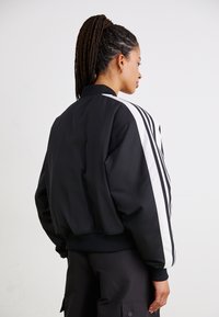 Svart bomberjakke med hvite striper, laget av glatt stoff. Har ribbestrikket krage og mansjetter, kort design og avslappet passform.