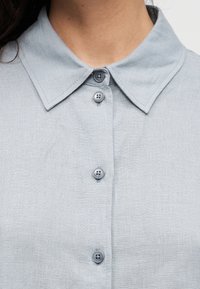 Lys blå linskjorte med spiss krage, seks grå knapper og en teksturert stoffoverflate. Enkel og klassisk design.