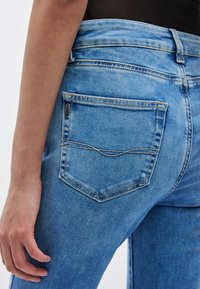 Ανοιχτό μπλε τζιν παντελόνι με πίσω τσέπη που διακοσμείται από ένα κυρτό ράψιμο και μια μικρή μεταλλική ταμπέλα· απαλή υφή, εφαρμοστό σχέδιο και μεσαία μέση.