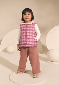 Polstret vest med pink og brun ternet mønster over en hvid langærmet skjorte. Løse brune sweatpants og bløde sneakers fuldender outfittet.