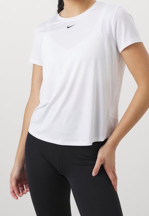 Femme portant un t-shirt Nike blanc uni à manches courtes et un legging noir, debout avec une main sur la hanche devant un fond blanc.