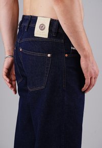 Dunkelblaue Denim-Jeans mit geradem Schnitt, Kupferrivets und einem Lederpatch am Bund, das Logo und Text zeigt.