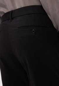 Gros plan sur un pantalon noir avec une poche arrière boutonnée et un logo Calvin Klein discret, porté avec une chemise claire.