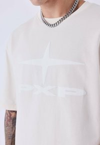 Crème kleurig t-shirt met wit grafisch logo "PXP" en sterrenontwerp. Gemaakt van katoen, heeft een ontspannen pasvorm en korte mouwen. Zilveren kettingaccessoire.