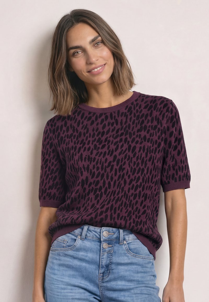 Femme souriante portant un pull violet à manches courtes avec des taches noires et un jean taille haute bleu clair, debout devant un fond uni.