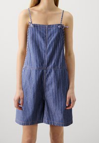 Sininen farkkuhaalarimallinen asu, jossa on valkoisia stripes, säädettävät hihnat, rypistetty kaula-aukko ja rento istuvuus shortsimuodossa.