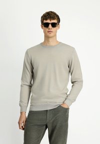 Młody mężczyzna w czarnych okularach przeciwsłonecznych, jasnoszarym swetrze i ciemnozielonych spodniach z kordurowej tkaniny, z jedną ręką w kieszeni, na białym tle.