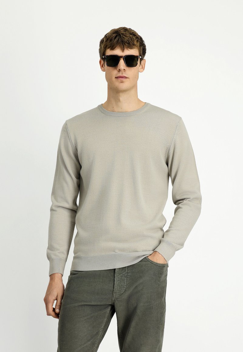 Młody mężczyzna w czarnych okularach przeciwsłonecznych, jasnoszarym swetrze i ciemnozielonych spodniach z kordurowej tkaniny, z jedną ręką w kieszeni, na białym tle.