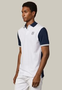 Homme portant un polo blanc avec des manches et un col bleu marine, arborant un petit logo "H" sur le côté gauche de la poitrine, debout de profil.