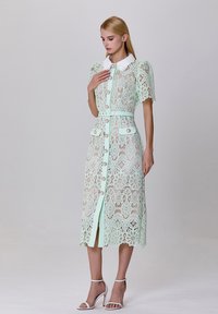 Robe en dentelle vert menthe avec un motif floral, manches courtes bouffantes, col blanc, fermeture à boutons sur le devant et deux poches avant.