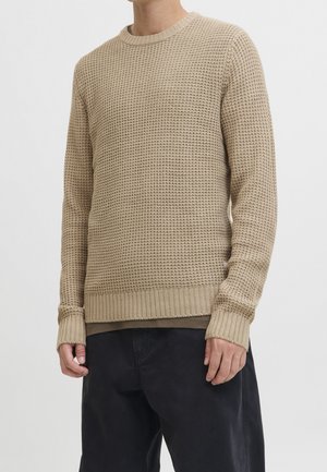 Pull en maille beige texturé avec poignets et ourlet côtelés porté sur un sous-vêtement marron, associé à un pantalon noir ample.