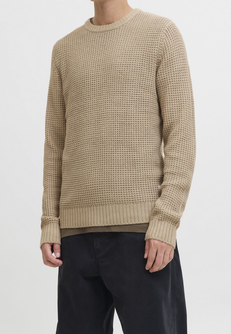 Pull en maille beige texturé avec poignets et ourlet côtelés porté sur un sous-vêtement marron, associé à un pantalon noir ample.