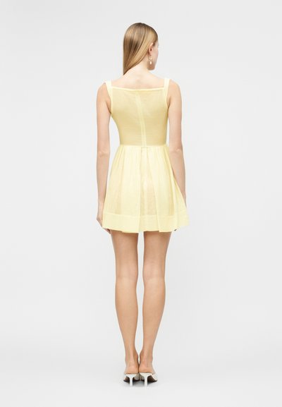 Γυναίκα με μακριά ξανθά μαλλιά, φοράει μινιμαλιστικό αμάνικο φούστα σε ανοιχτό κίτρινο χρώμα και λευκά παπούτσια με ψηλό τακούνι, στέκεται με την πλάτη στην κάμερα.