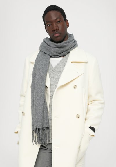 Sciarpa grigia drappeggiata attorno al collo, indossata sopra un maglione grigio chiaro a maglia e un cappotto bianco doppiopetto, con una texture liscia e estremità frangiate.