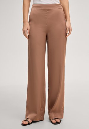 Femme portant un pantalon marron taille haute à jambes larges, un haut blanc et des sandales noires à bout ouvert, debout devant un fond uni.