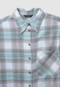 Camicia a quadri blu chiaro, grigia e bianca con bottoni. Presenta un colletto, taschino sul petto e bottoni marroni con una texture del tessuto morbida.