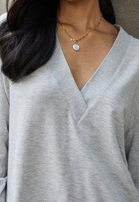 Sweat-shirt gris clair en v avec une texture douce. Les accessoires comprennent un collier en chaîne dorée avec un pendentif rond en pierre blanche.