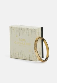 Gold-tone bangle bracelet features three clear crystals and a design with round cutouts. Accompanied by a decorative cream box with gold accents.