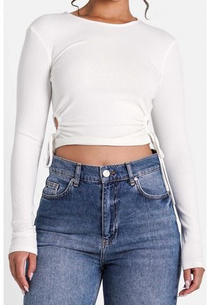 Frau trägt ein weißes, geripptes Langarm-Crop-Top mit seitlichen Kordelzügen und hoch taillierte blaue Jeans.