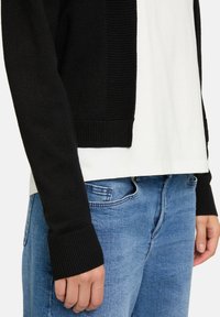 Svart stickad cardigan med ribbad textur, kort design och långa ärmar, parat med en vit t-shirt och ljusblå jeans.