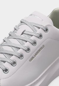 Άσπρο sneaker Karl Lagerfeld με γκρι κορδόνια με σχέδιο, ασημένιες τρύπες και ανάγλυφο όνομα της μάρκας στο πλάι και στη γλώσσα.