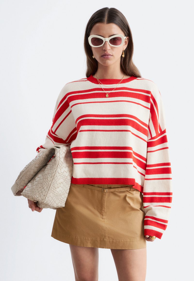 Femme aux longs cheveux foncés portant des lunettes de soleil blanches, un pull rayé rouge et blanc, une mini-jupe beige, tenant un grand sac tressé beige.
