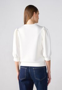 White pullover with puff sleeves, rounded neckline, and ribbed cuffs. Paired with dark blue jeans featuring back pockets and stitching details.