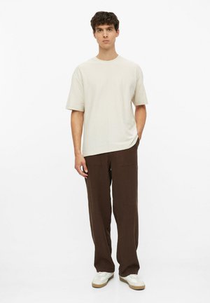 Junger Mann, der ein schlichtes beige kurzärmeliges Hemd, lockere braune Hose und weiße Sneakers trägt, vor weißem Hintergrund.