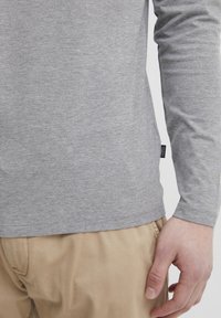 Camisa de manga larga gris hecha de una tela suave, con una etiqueta en la costura lateral. Enfocándose en la textura lisa y el diseño ajustado. Combinada con pantalones beige.