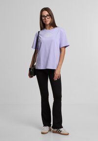 Jauna moteris su ilgesniais rudais plaukais, dėvinti juodus akinius, levandų spalvos T-shirt, juodas kelnes, baltus sportbačius ir nešanti juodą per petį dėklo krepšį.