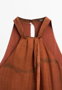 Rust-colored halter top with a keyhole back, featuring a gathered front and a tie detail. Button closure at the neck. Fashion item.