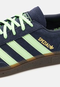 Marinblå mockaskor med gröna accenter, som har tre horisontella ränder, platta skosnören och gul "SPEZIAL"-logotyp på sidan.