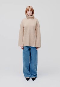 Pullover beige à col roulé côtelé, coupe oversize, associé à un jean bleu ample et des escarpins noirs à talons pointus. Design simple et épuré.