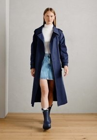 Cappotto trench blu navy con ampio colletto, abbinato a un dolcevita bianco, gonna di jeans e stivali di gomma neri, su uno sfondo neutro.
