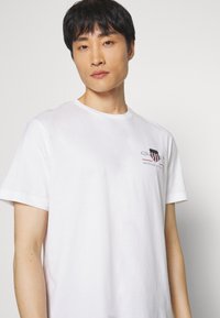 T-shirt en coton blanc avec un col rond et des manches courtes. Présente un petit logo avec des accents rouges, bleus et noirs sur le devant.