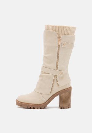 Botte beige mi-mollet à talon avec fermeture éclair latérale, détails à bride, manchette côtelée en tricot, talon bloc marron texturé et semelle.