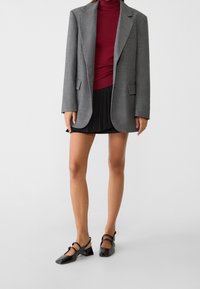 Veston à carreaux gris, col roulé bordeaux ajusté, mini-jupe noire plissée, et chaussures à talons carrés en cuir verni noir avec brides à la cheville.