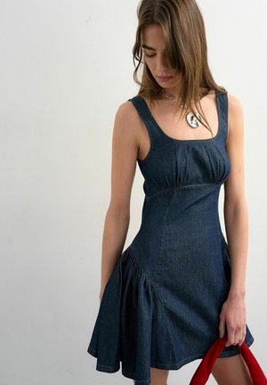 Vrouw in mouwloze donkerdenim jurk die een rood stoffen voorwerp vasthoudt, omlaag kijkt, een zilveren hangerketting draagt tegen een effen achtergrond.