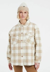 Vaatteena on beigellä ja valkoisella ruutukuviolla varustettu tikattu takki, jossa on kaksi läpällistä taskua, napit ja kauluspanta. Takki on yhdistetty beigessä olevan myssyn kanssa.