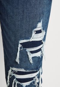 Jean en denim bleu foncé avec de grandes déchirures horizontales laissant voir des fils blancs effilochés et un tissu bleu plus clair en dessous.