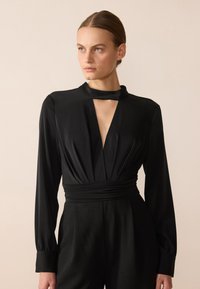 Combinaison noire avec des manches longues, un décolleté en V profond et une taille drapée. Fabriquée dans un tissu doux et lisse avec un éclat subtil.