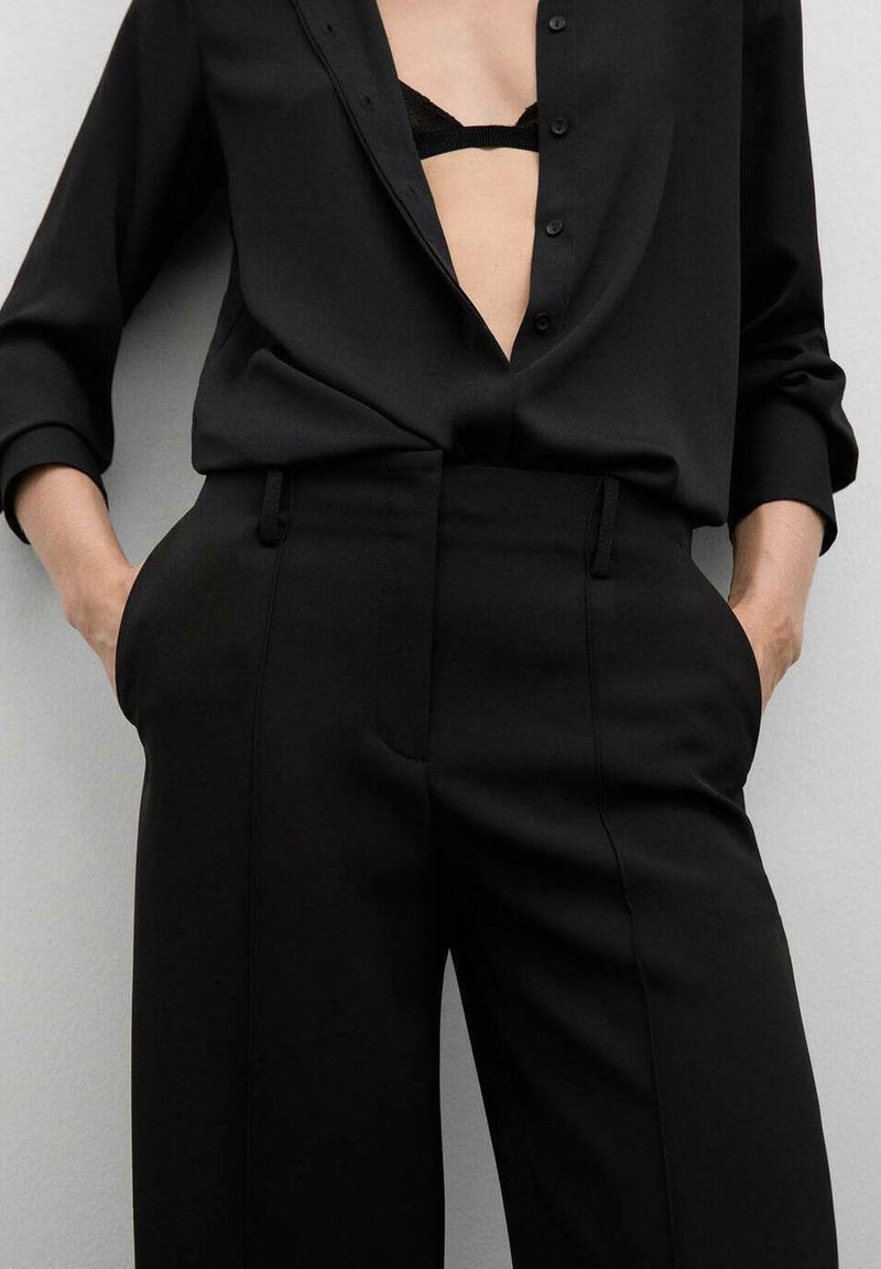 Pantalones negros a medida y camisa de mangas largas a juego. La camisa presenta un escote en V profundo con botones y una textura suave. Los pantalones tienen bolsillos laterales.