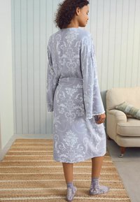 Robe floreale grigio chiaro con maniche lunghe e vita con cravatta, con bordo in pizzo e calzini grigi abbinati, posata su un tappeto testurizzato.