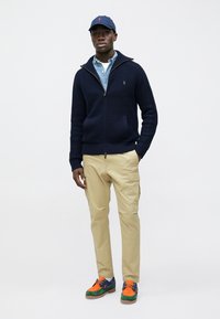 Jersey de punto azul marino con cremallera, camisa azul claro, pantalones cargo caqui y zapatillas multicolor con detalles en verde, naranja y azul.