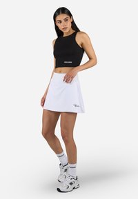 Must mustriba crop top musta "PEGADOR" valges trükituna, koos valgete lühikeste seelikutega, millel on diskreetne logo. Spordisokid ja tennised lõpetavad välimuse.