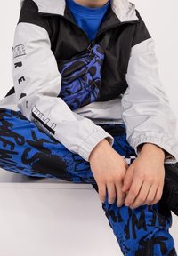 Kurtka w stylu color-block w czerni i bieli, z graficzną torbą na ramię. Niebieskie spodnie z wyraźnym czarnym napisem i wzorem rozprysków.